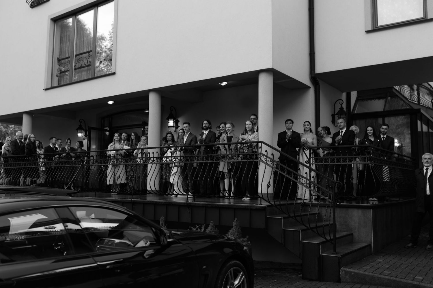 A black and white photo shows a crowd of people gathering on balconies above a parked car at an event.