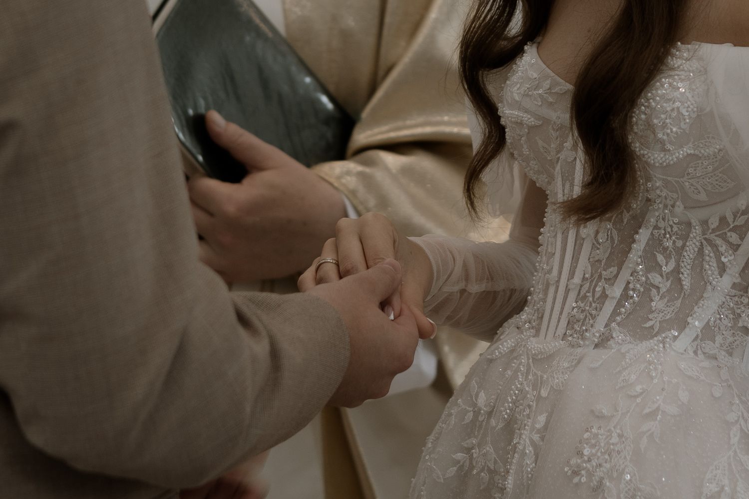 A couple holds hands during a wedding ceremony, with the bride wearing an intricate lace dress and beige tones.