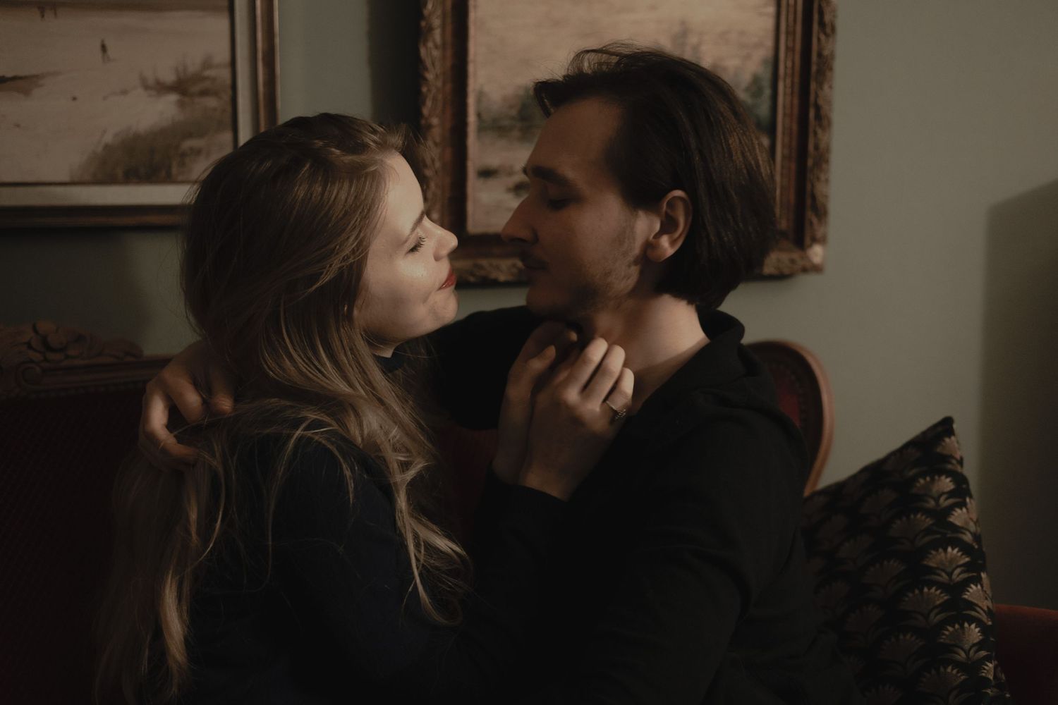 A romantic couple shares an intimate moment in dim lighting with vintage framed artwork on the wall behind them.