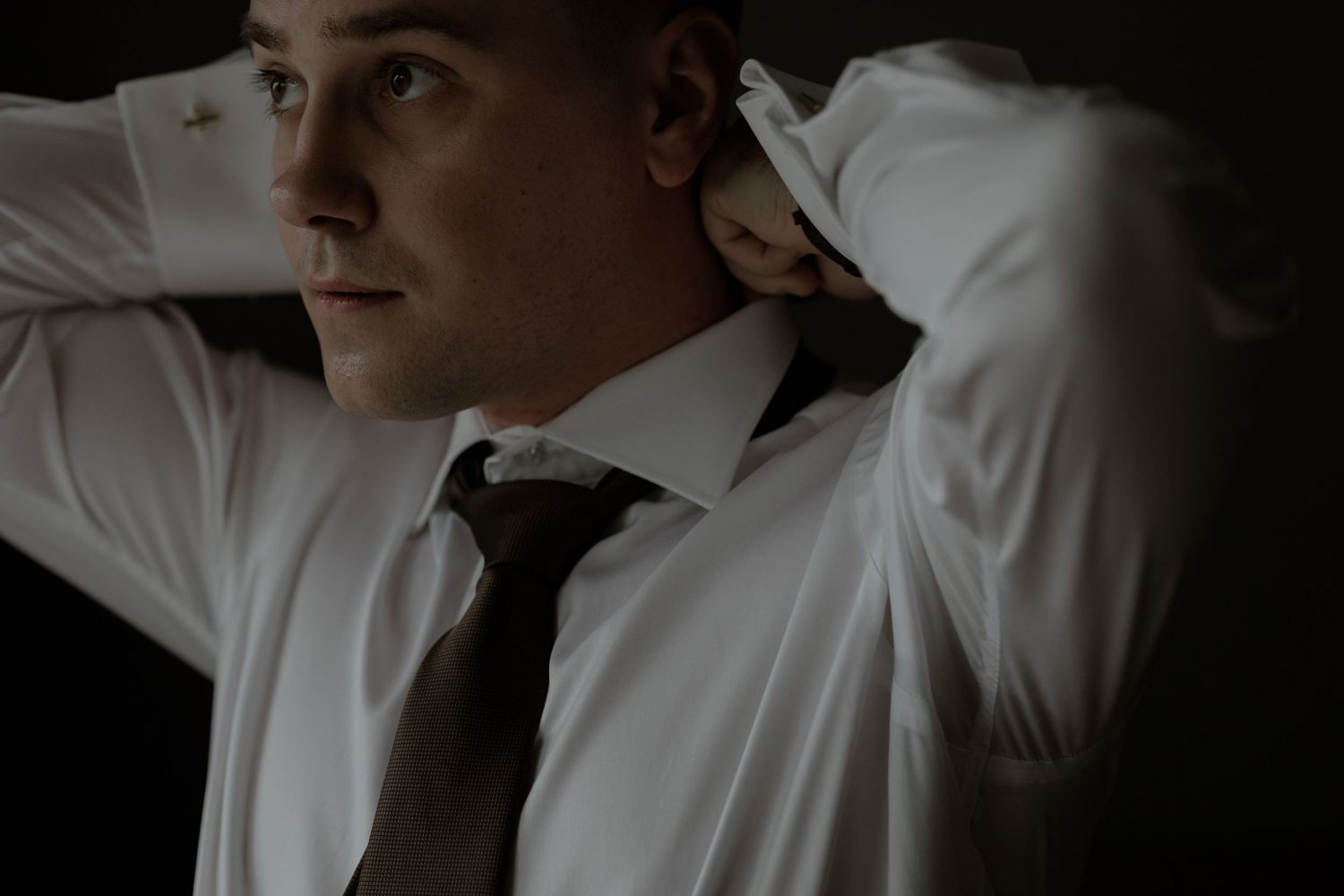 Moody portrait shot in dark lighting of someone wearing a white dress shirt and dark tie with hands behind their head.