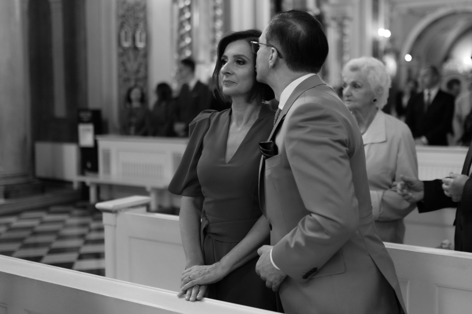 A couple stands together in a church pew during a formal gathering, captured in black and white.
