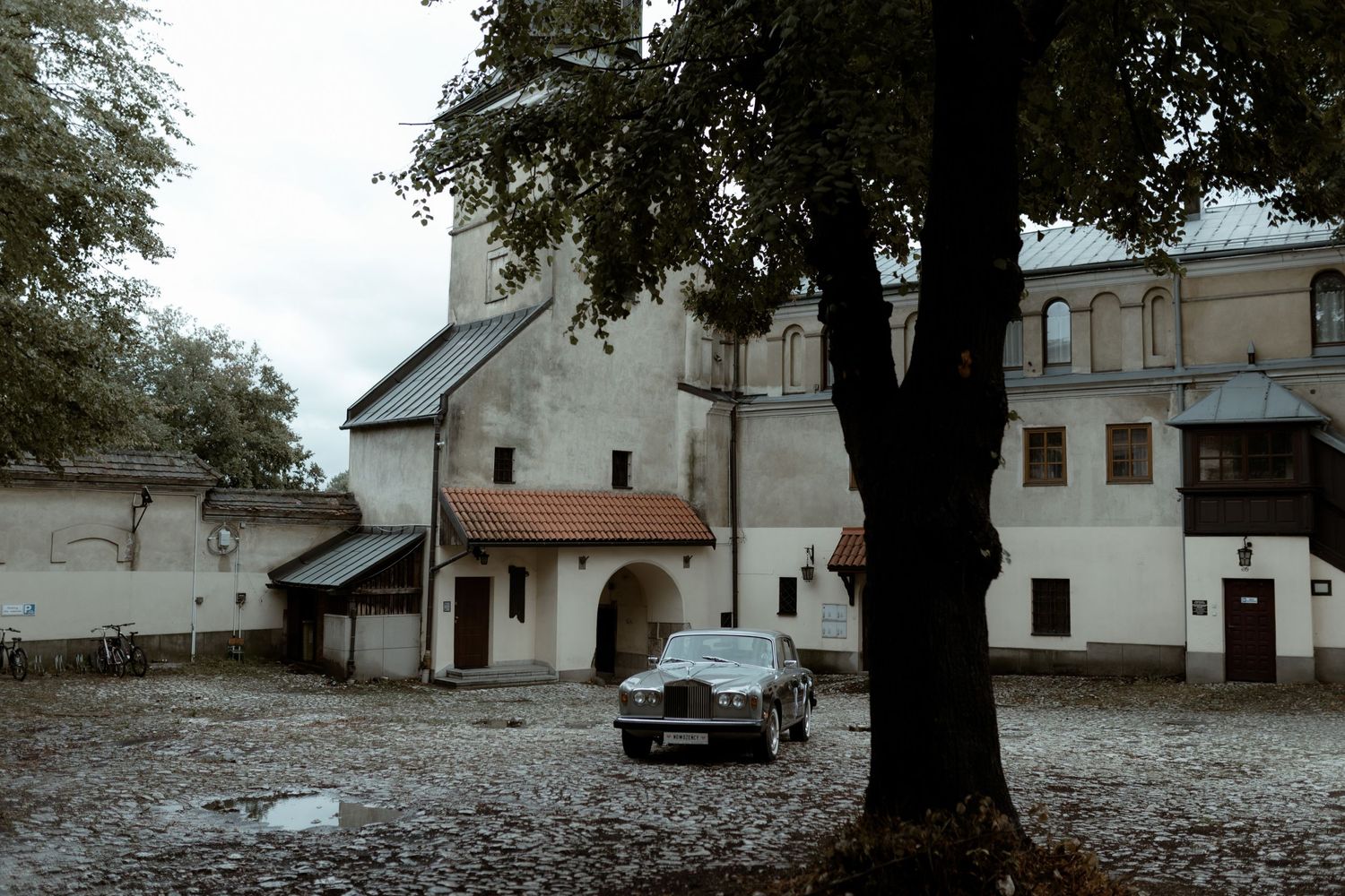 A vintage car parked in a courtyard of an old white church building with a dark tree in the foreground.