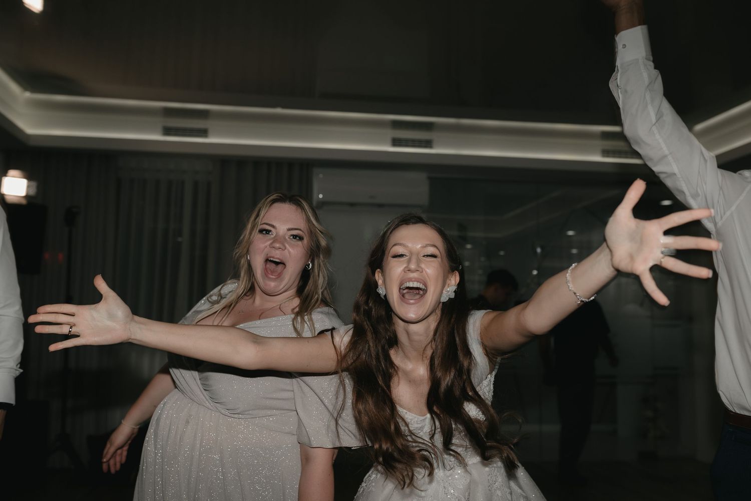 Two wedding guests dancing joyfully with arms outstretched on a dark dance floor.