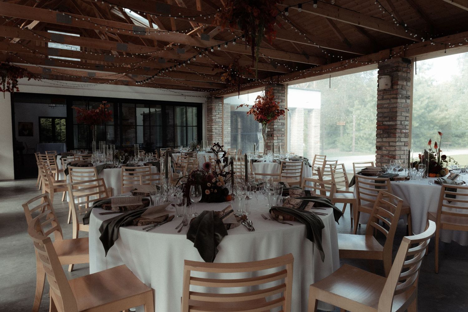 Wedding reception venue with rustic exposed brick walls, wooden chairs and elegant round tables set with white linens.