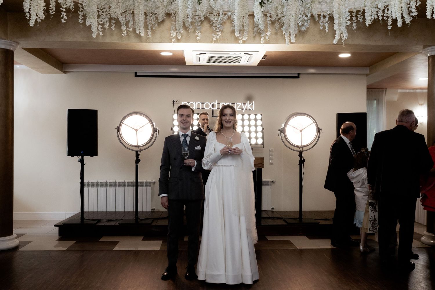 A couple stands together at their wedding reception with decorative clocks on the wall and crystal chandeliers above.