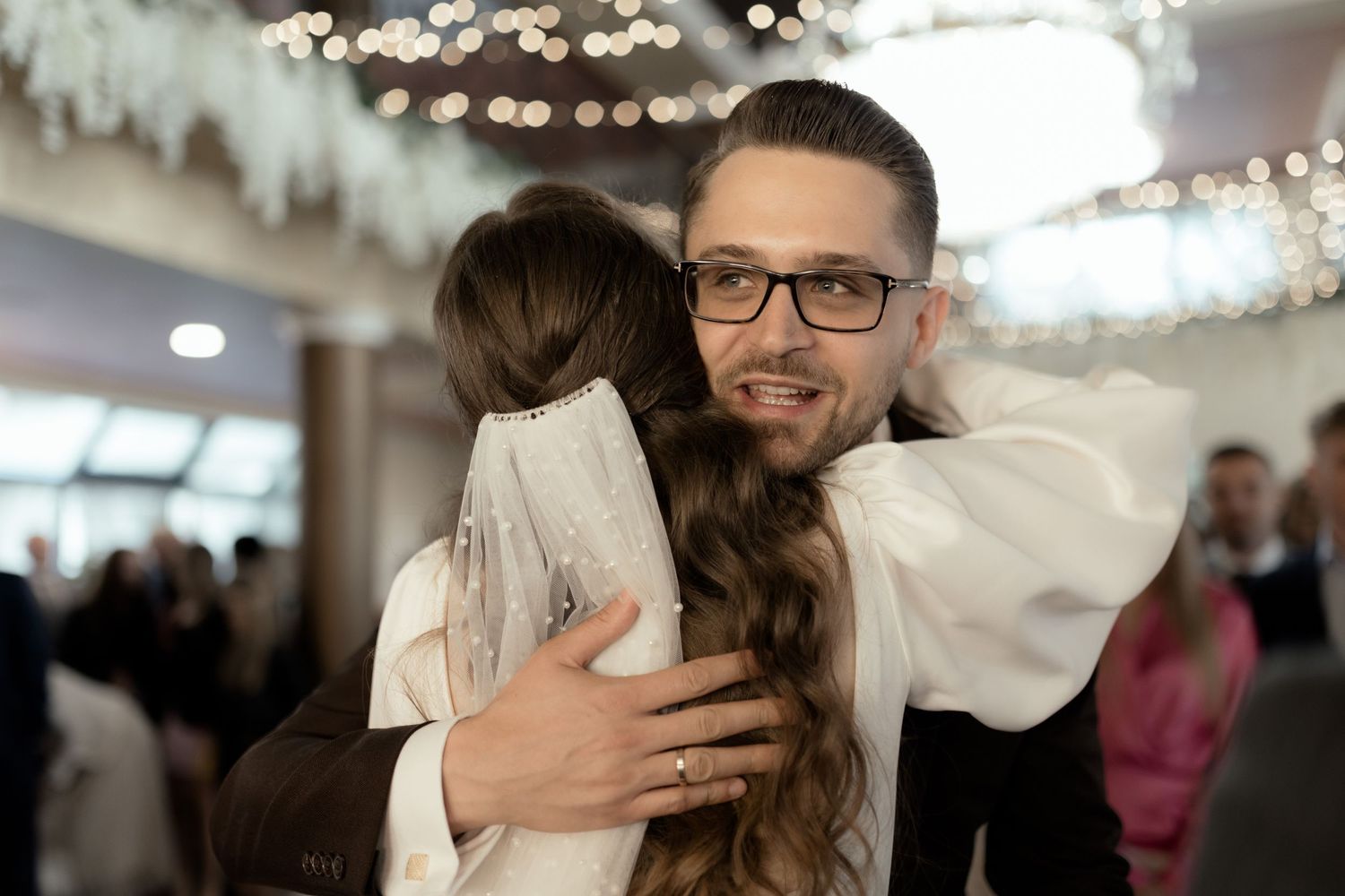 Two people share an emotional embrace at an indoor event with twinkling lights in the background.
