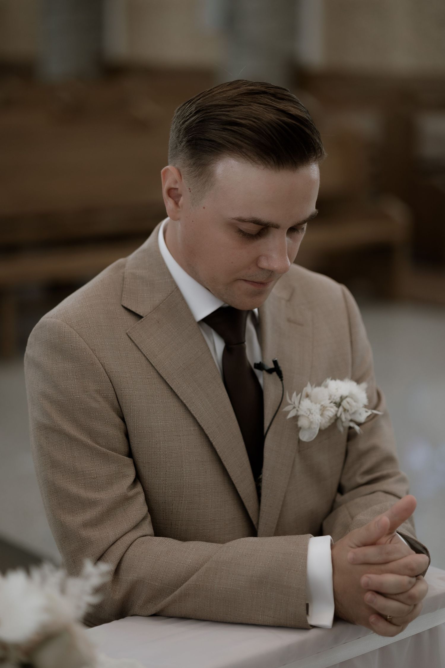 A groom in a tan suit and black tie looks down thoughtfully while wearing a white floral boutonniere.