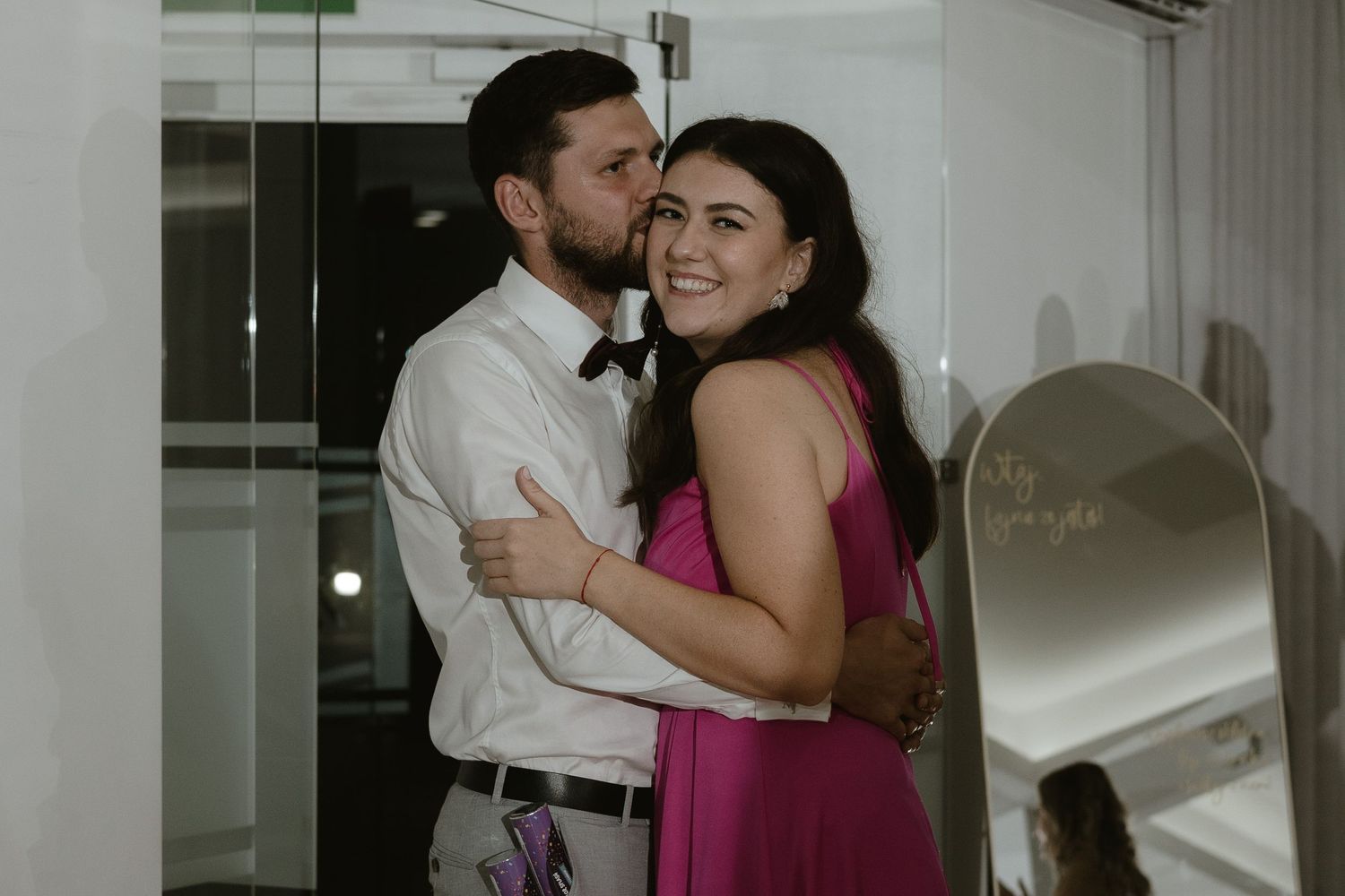 A couple embraces while dancing together at an indoor venue, with one person wearing a bright pink dress.