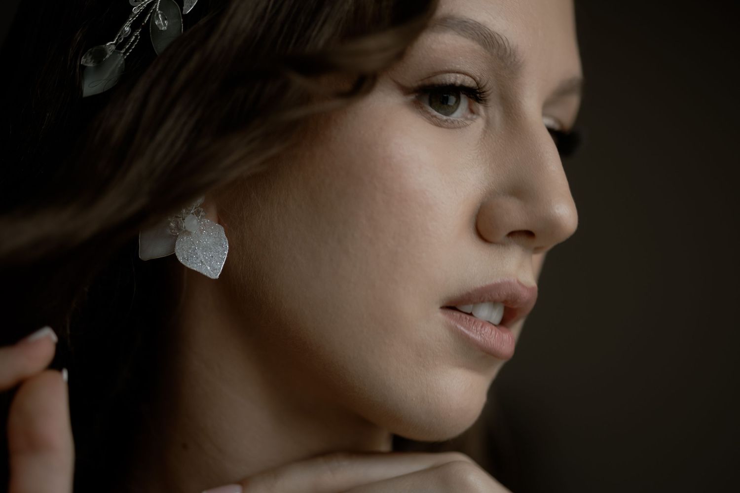 Side profile portrait of someone with delicate pearl earrings and soft natural makeup against dark background.
