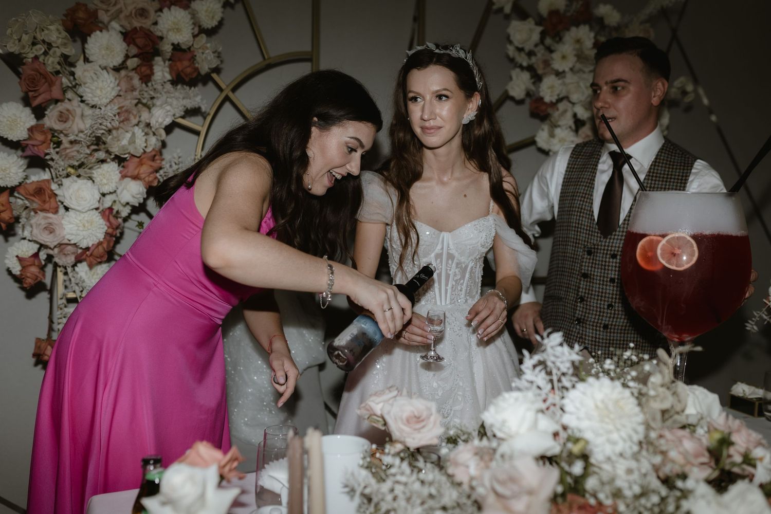 Wedding reception scene with guests in formal attire gathered around floral-decorated table at evening celebration.