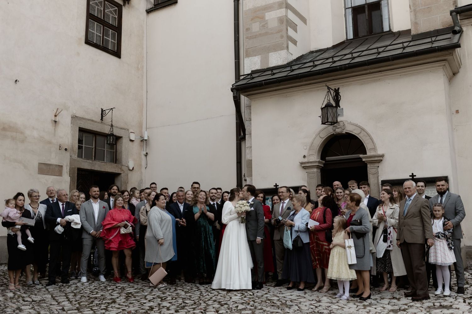 Wedding guests gather outside a historic church with white walls and arched doorway on a cobblestone plaza.