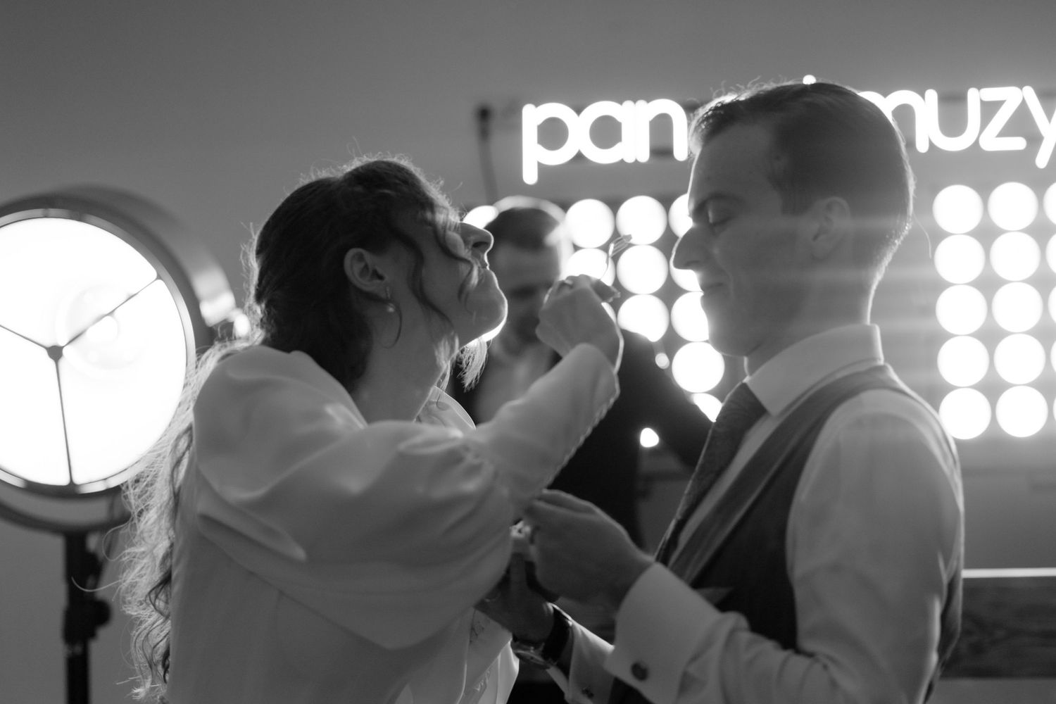 Black and white photo of people singing into a microphone at a party venue with illuminated lights in the background.