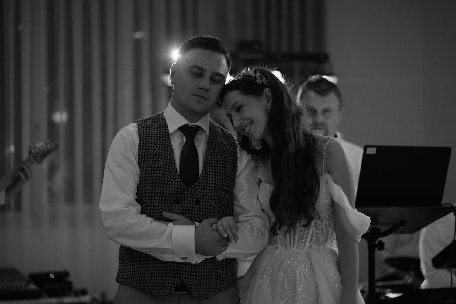 A couple dances intimately at their wedding reception, with blurred musical equipment in the background.
