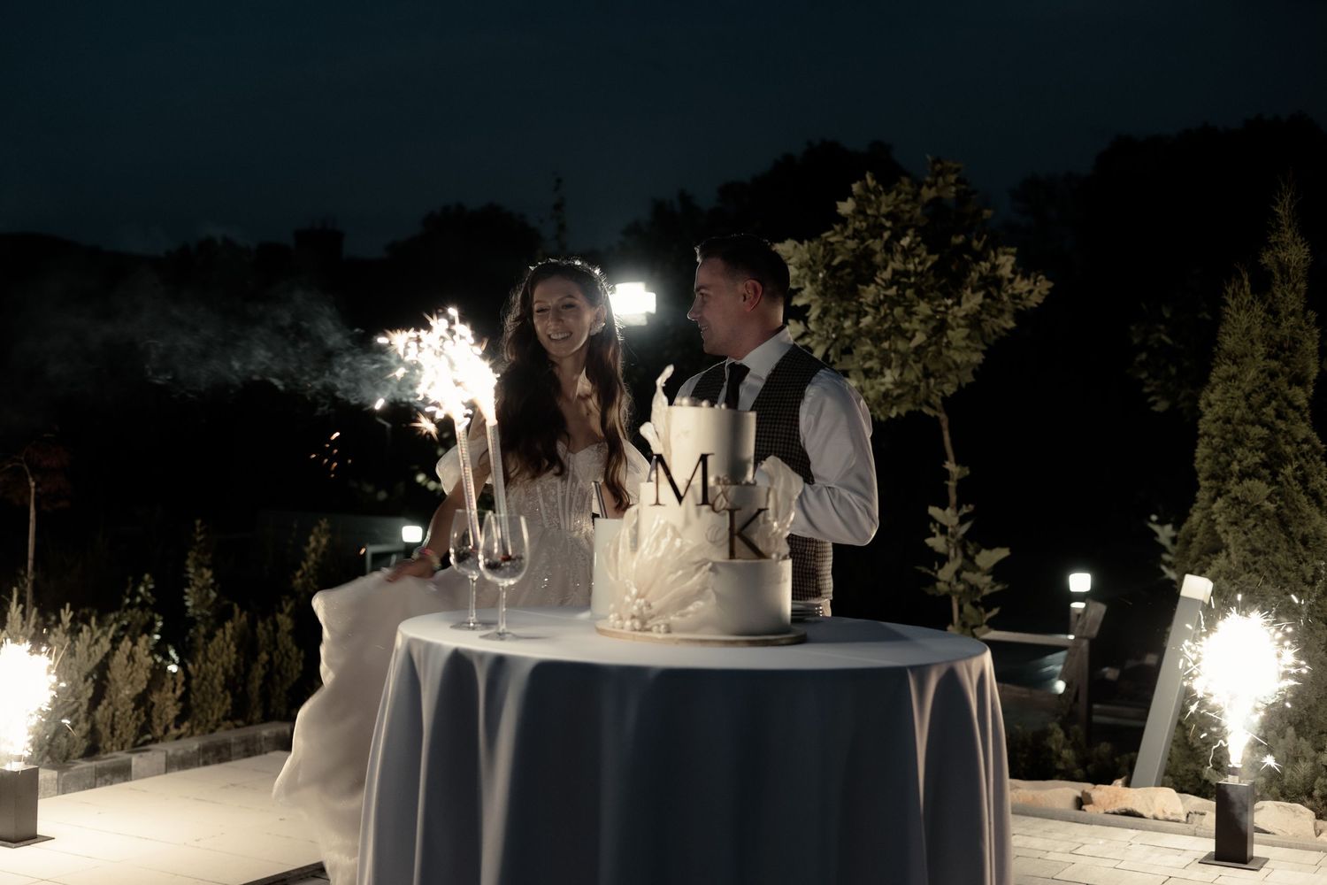 A couple cuts their elegant white wedding cake at an outdoor evening reception under twinkling lights.