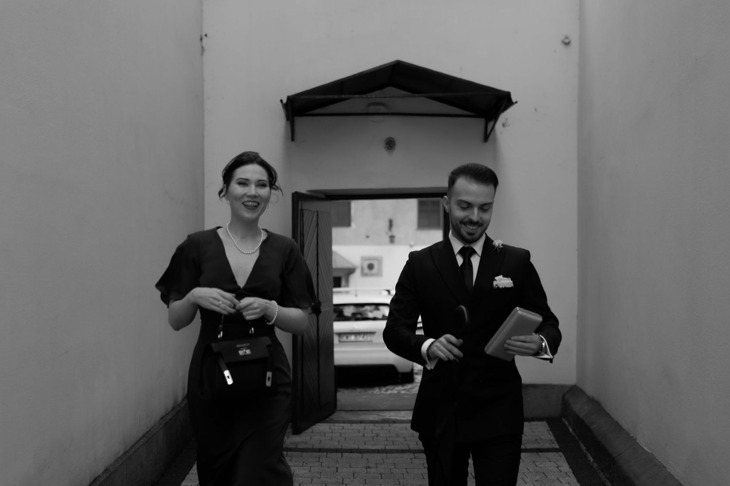 A black and white photograph shows two people smiling as they walk together through a hallway with an arched entrance.