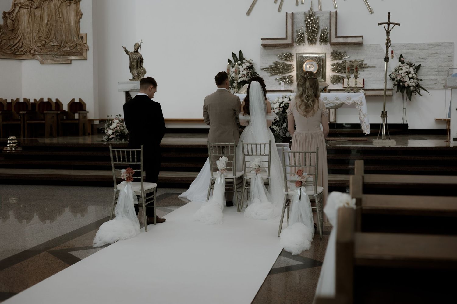 A traditional Catholic wedding ceremony in progress inside a church sanctuary with white aisle runner and floral decorations.