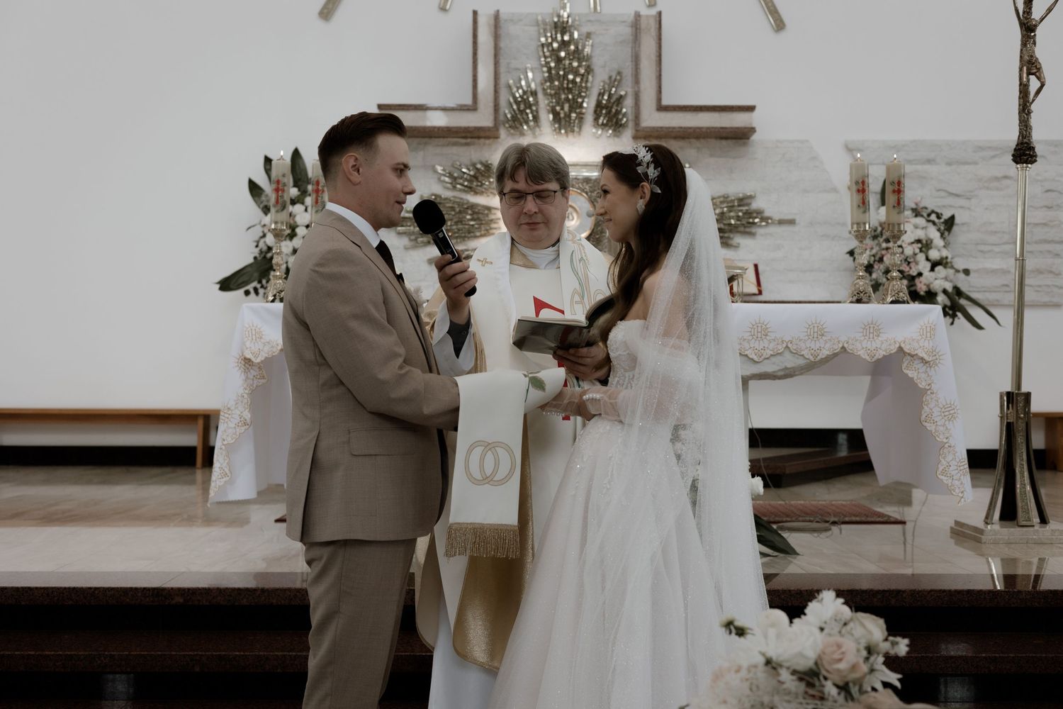 A couple stands at the altar during their wedding ceremony, with an officiant leading the service in a light-filled church.