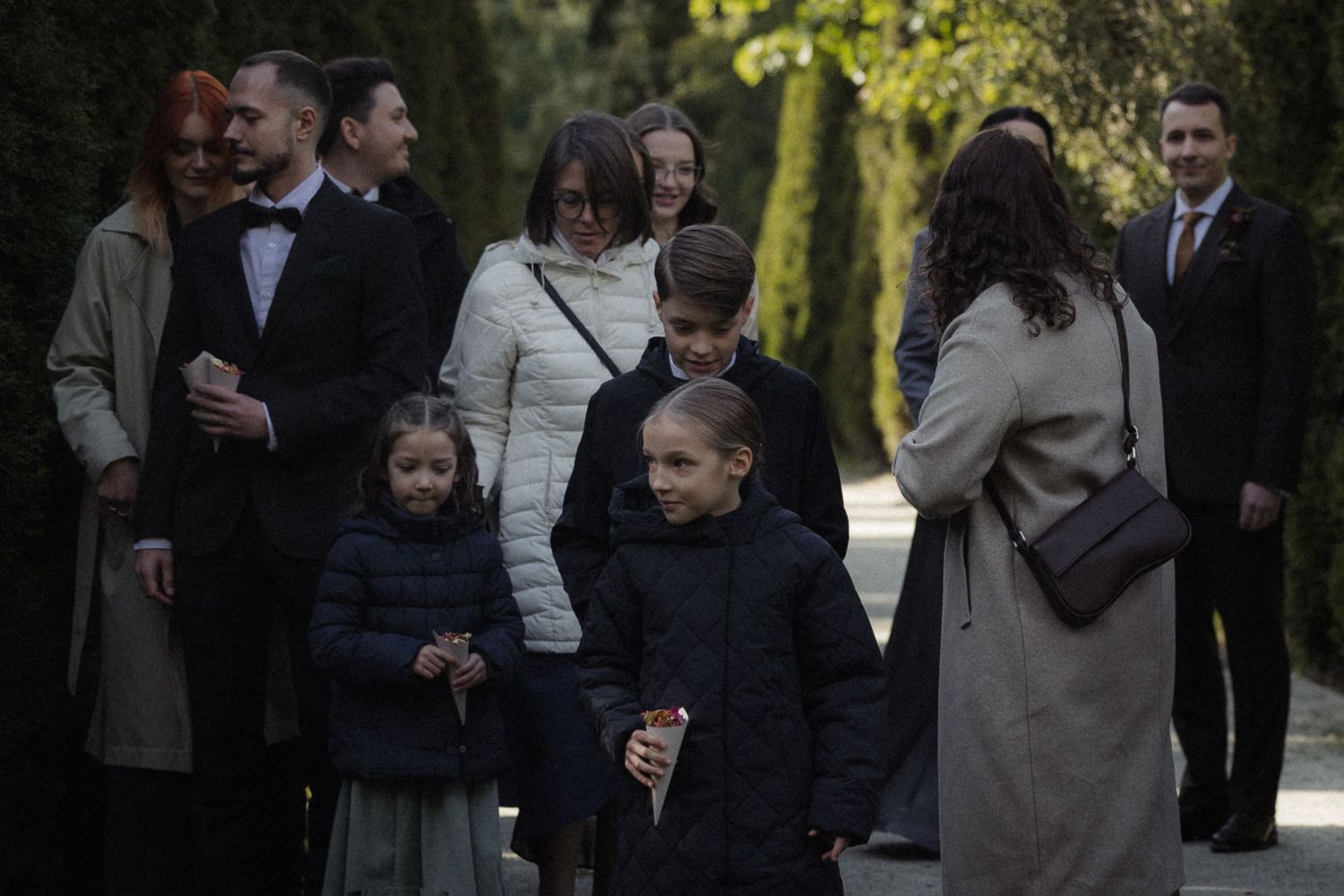 A group of people in dark coats and formal attire gather outdoors on a tree-lined path during daytime.