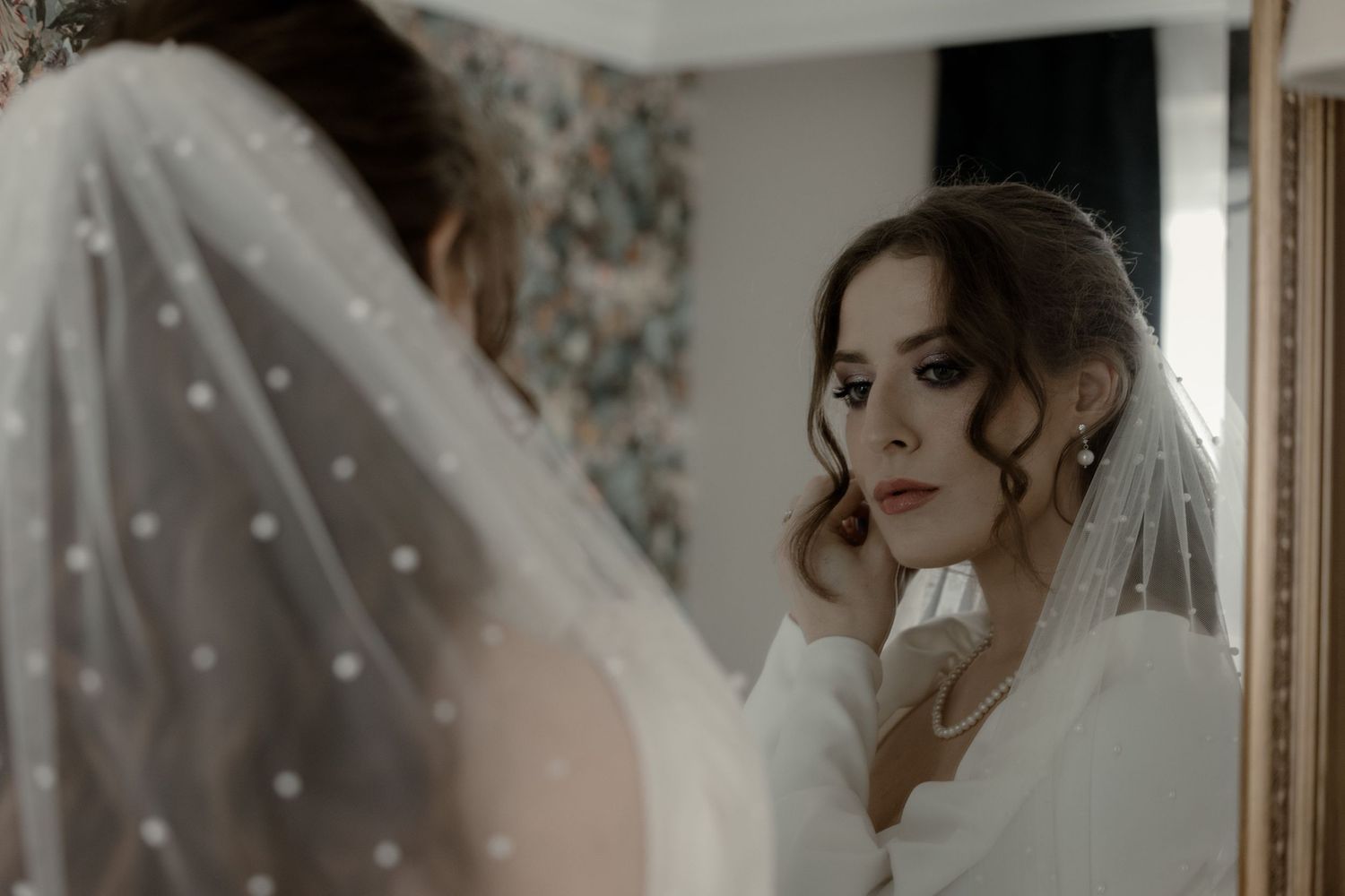 A bride wearing a white wedding dress and veil gazes thoughtfully at her reflection in a mirror on her wedding day.