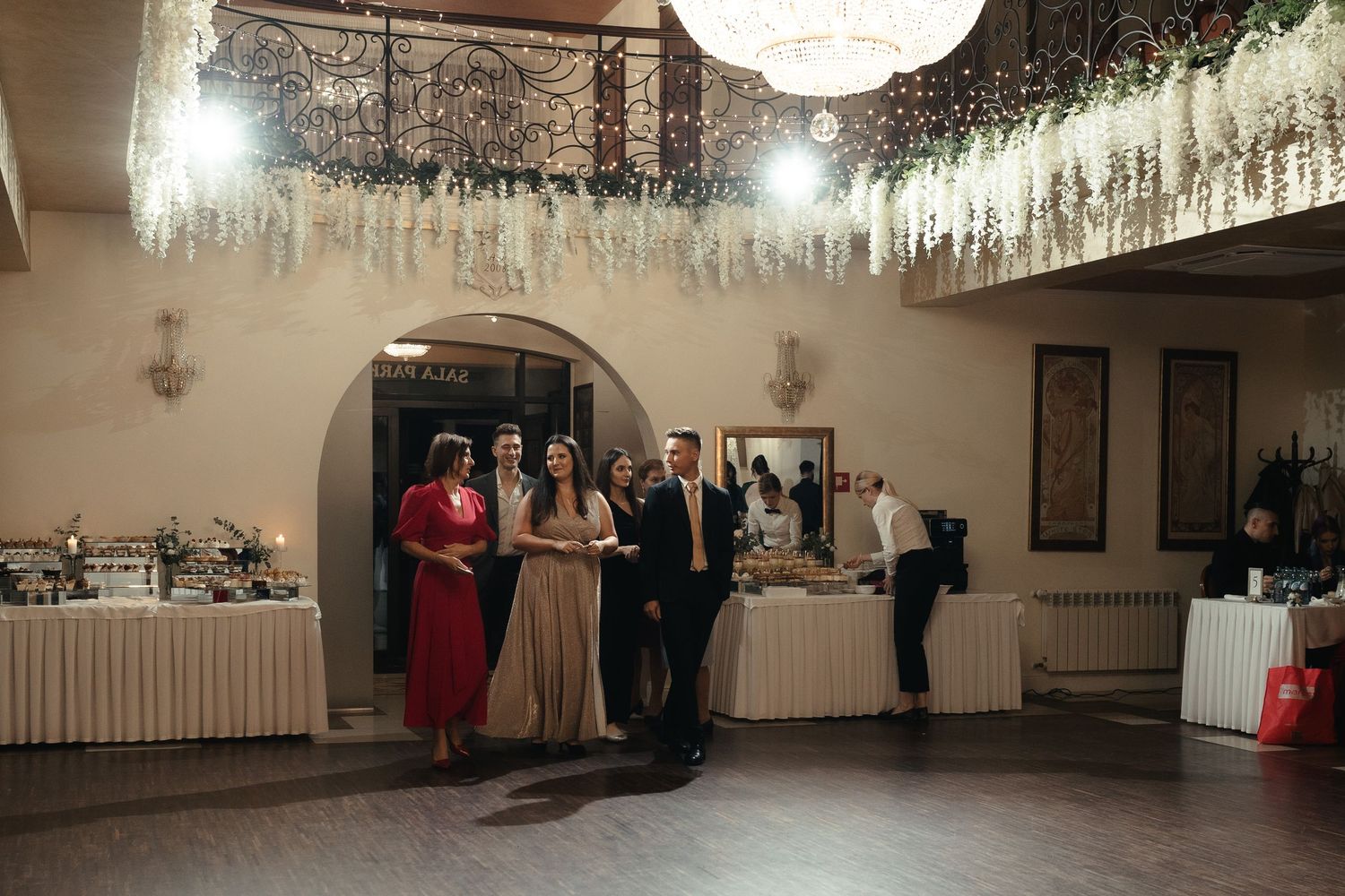 Wedding guests stand in an elegant reception hall decorated with hanging crystal lights and a buffet table along the wall.