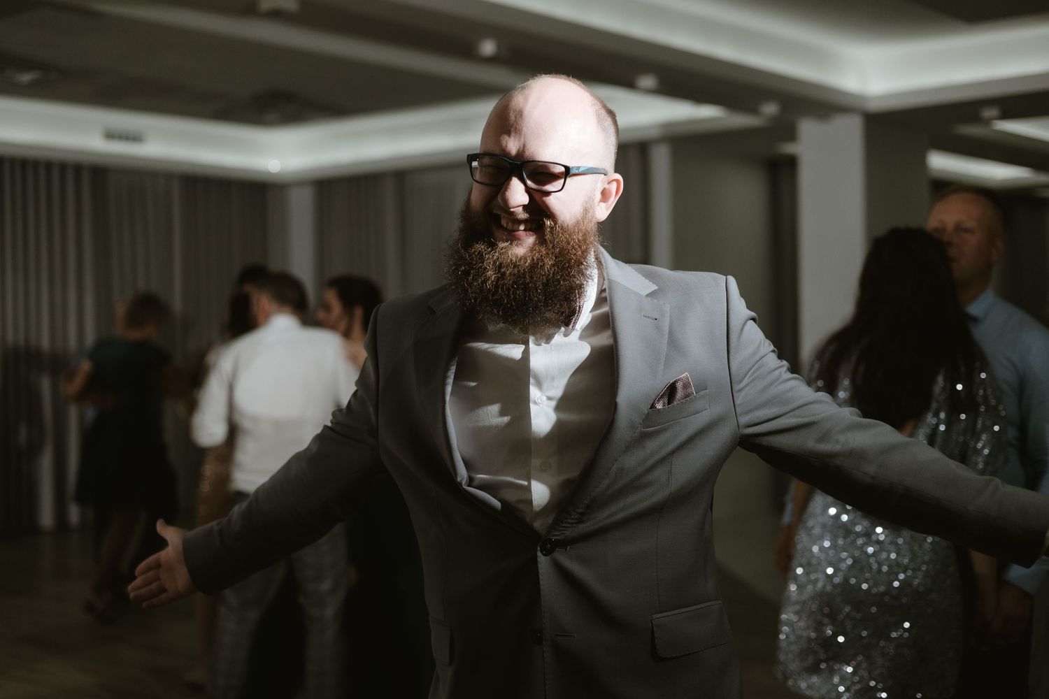 A bearded man in a grey suit dances enthusiastically at an indoor celebration.