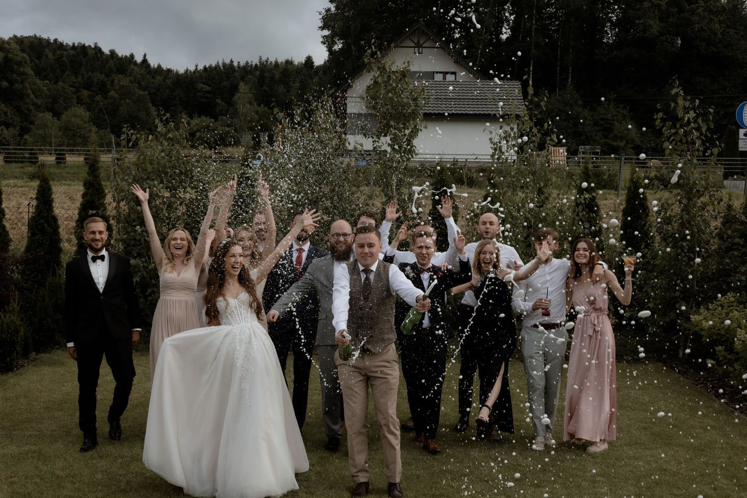 Joyful group celebrating on a grassy lawn with confetti flying through the air at an outdoor countryside wedding.