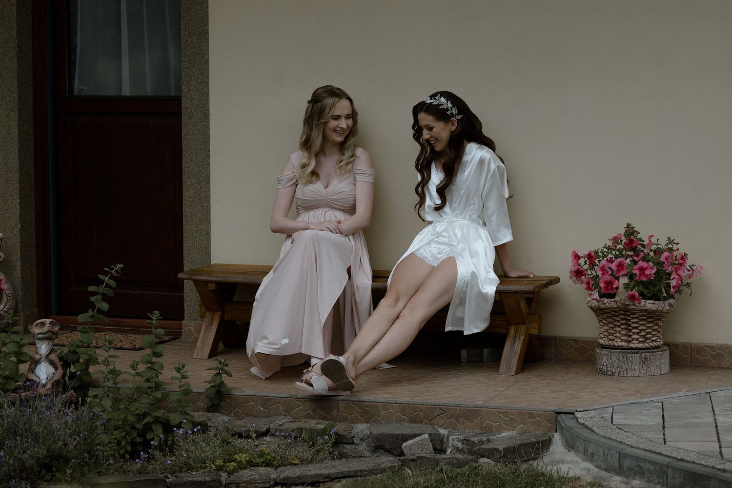 Two people in romantic dresses sit together on rustic wooden porch steps beside pink potted flowers.