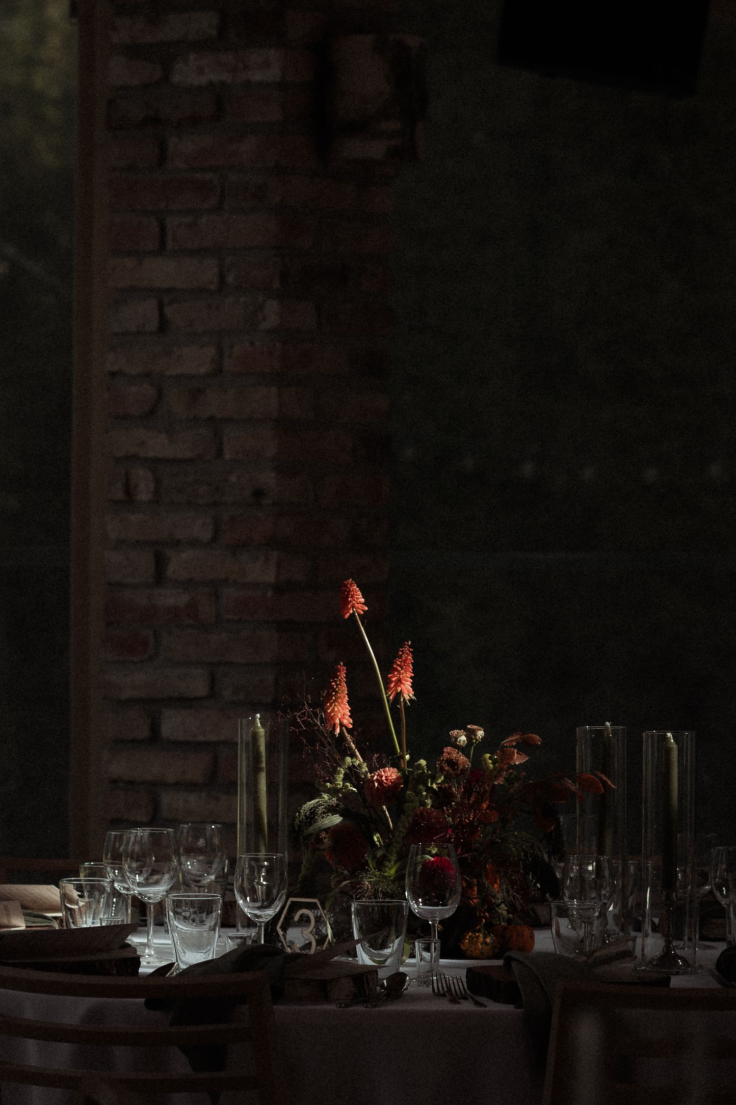 A dark and moody table setting with wine glasses and red floral centerpieces against a rustic brick wall.