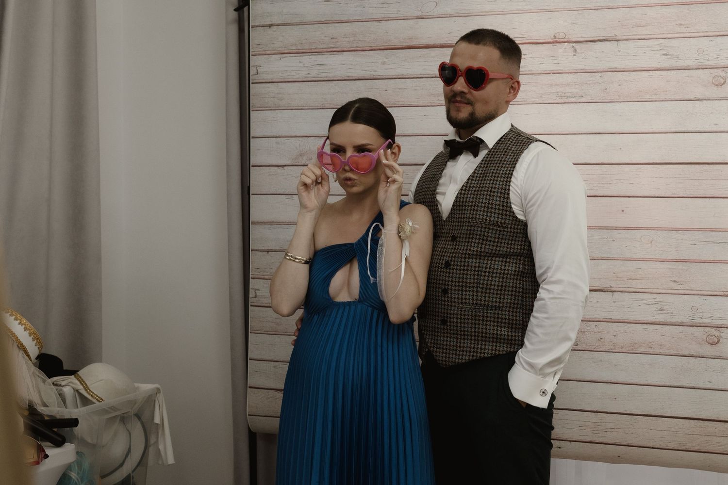 A couple poses in front of a wooden wall wearing sunglasses, a blue dress and formal vest attire.