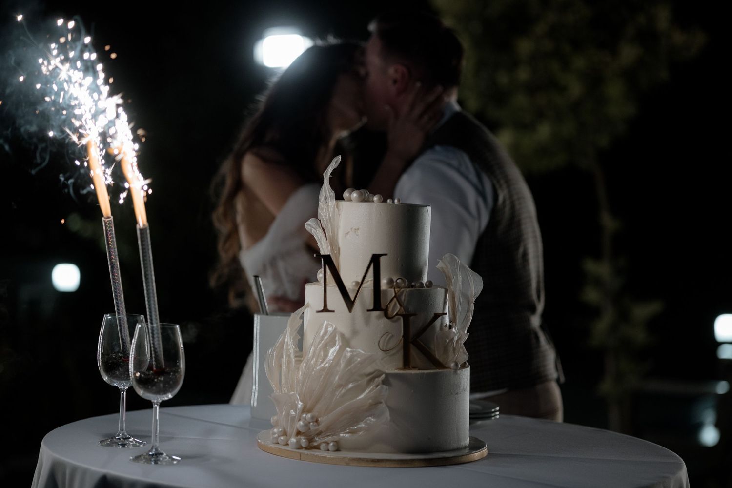 An elegant white wedding cake with M and K monogram sits on a table with sparklers and champagne flutes at a nighttime reception.