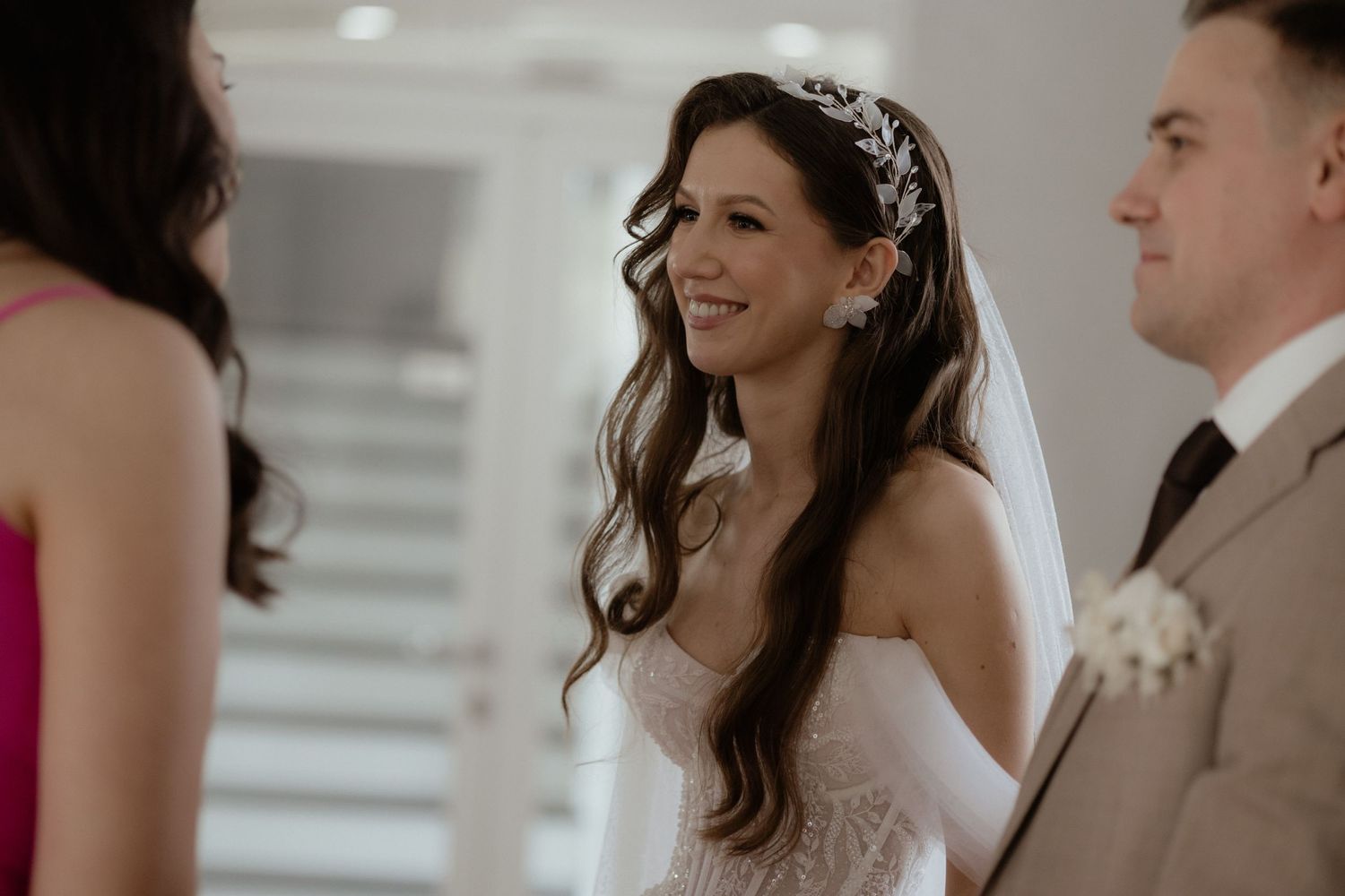 A bride smiles joyfully during her wedding ceremony while wearing a strapless white gown and long wavy hair.