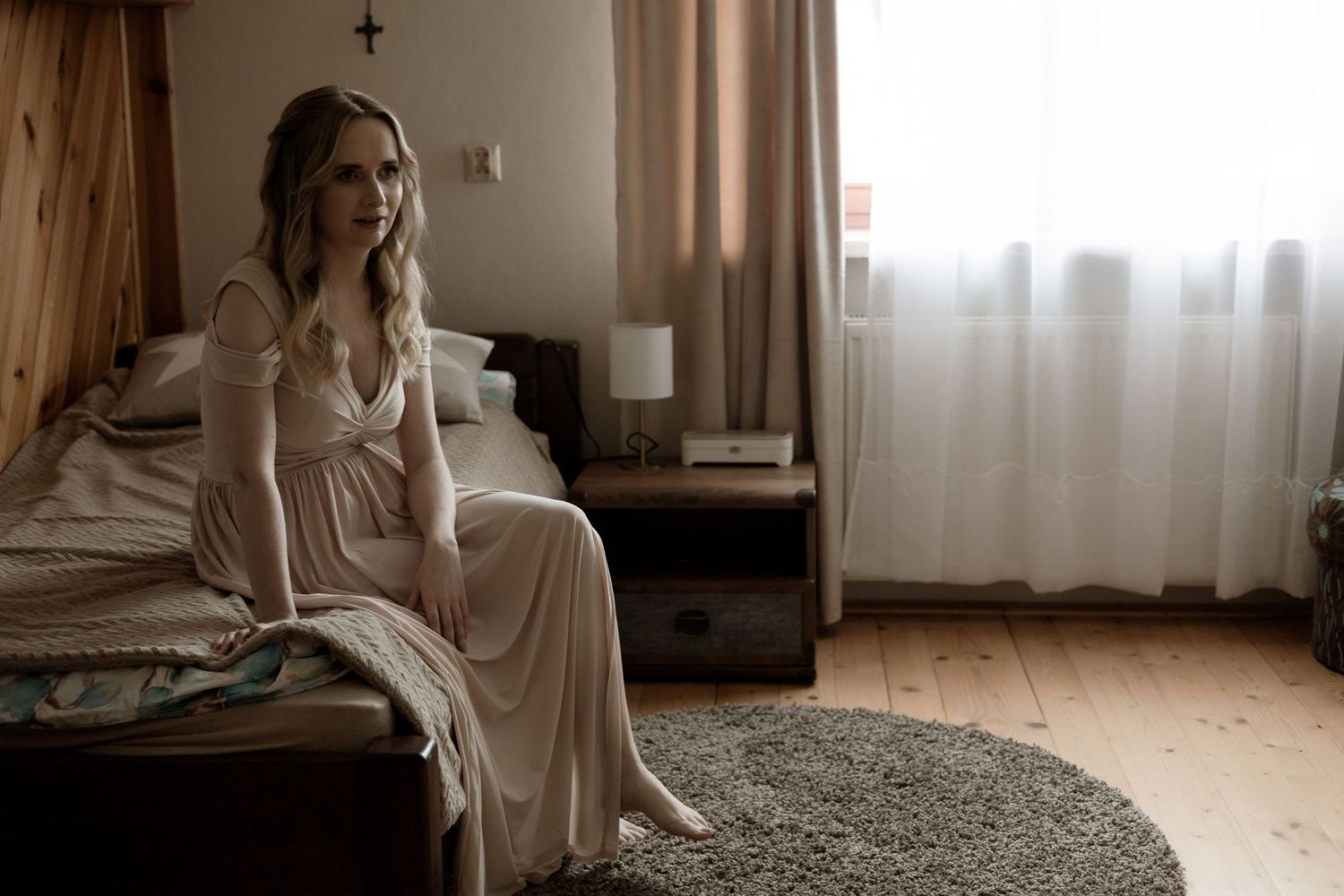 A woman in a flowy white dress sits pensively on the edge of a bed in a sunlit bedroom.