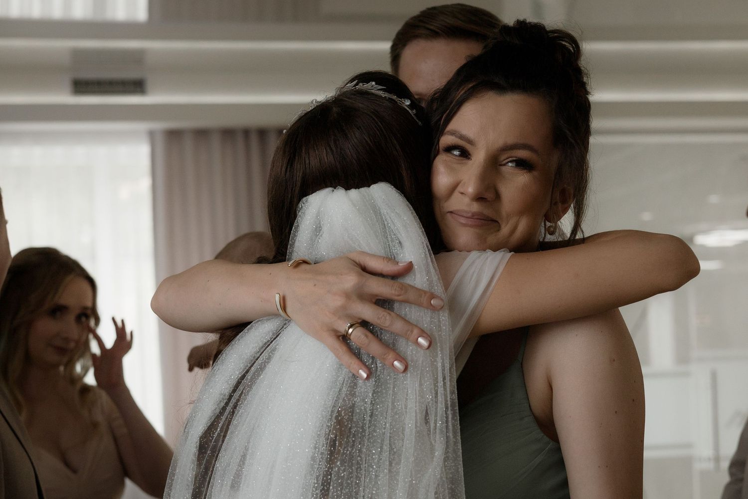 Two people share a warm embrace at an indoor gathering while another person looks on in the background.