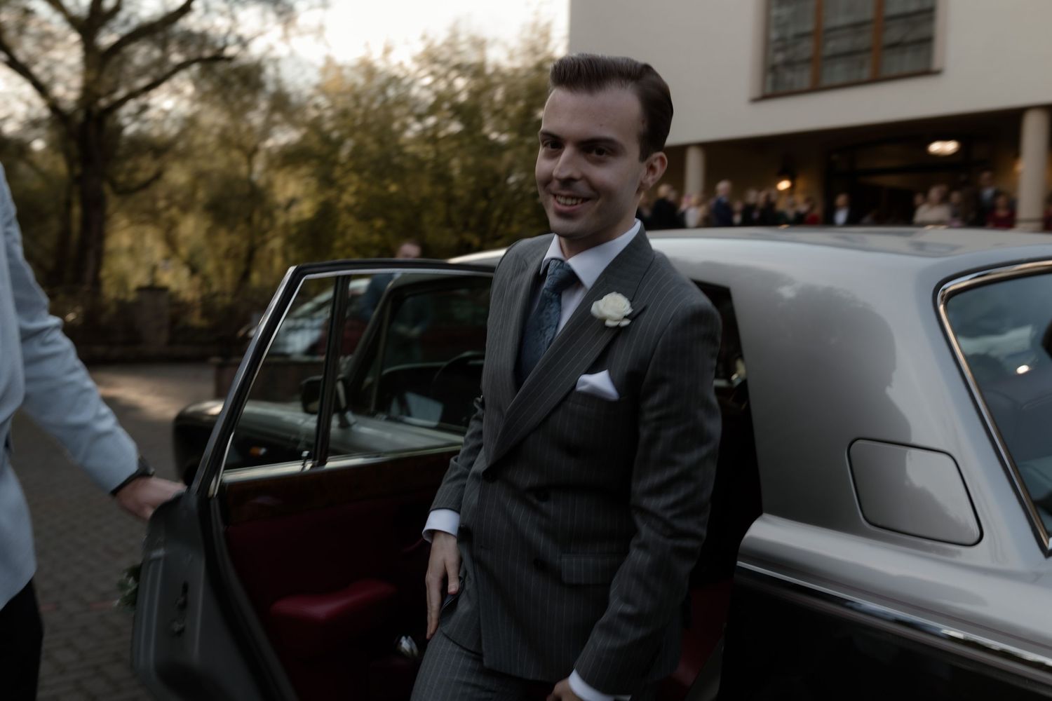 A well-dressed person in a dark gray suit and boutonniere stands beside a vintage car on an evening.