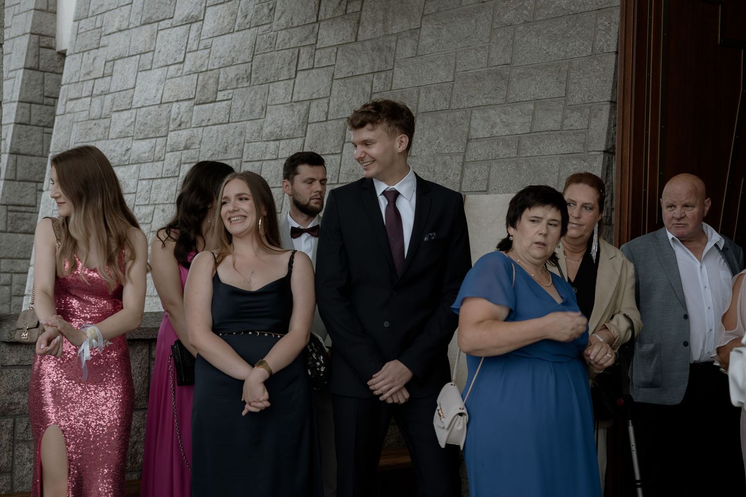 A group of people dressed in formal attire gathering outside a stone church or building entrance.