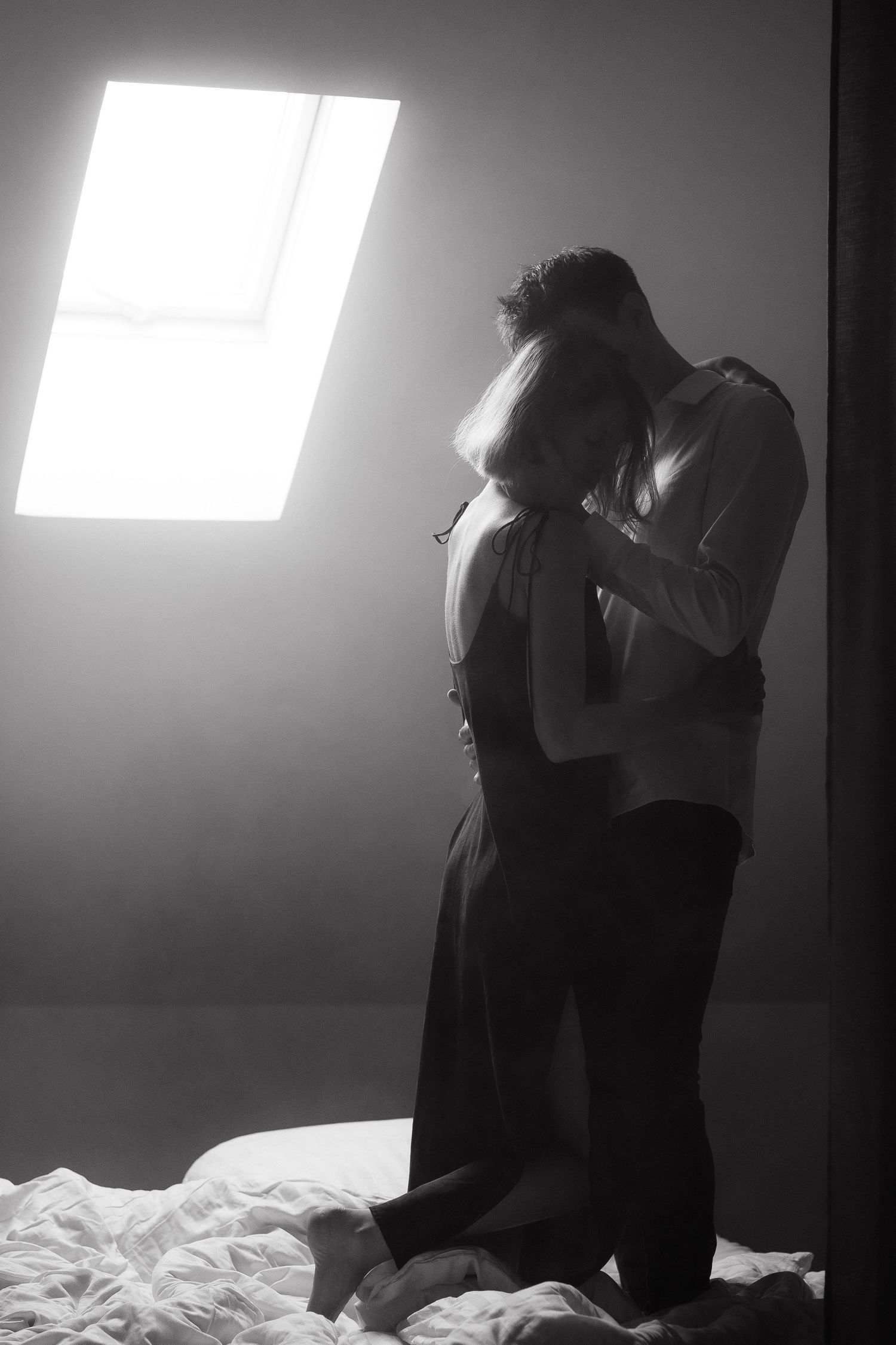 Silhouetted couple in an intimate embrace near a bright window in black and white.