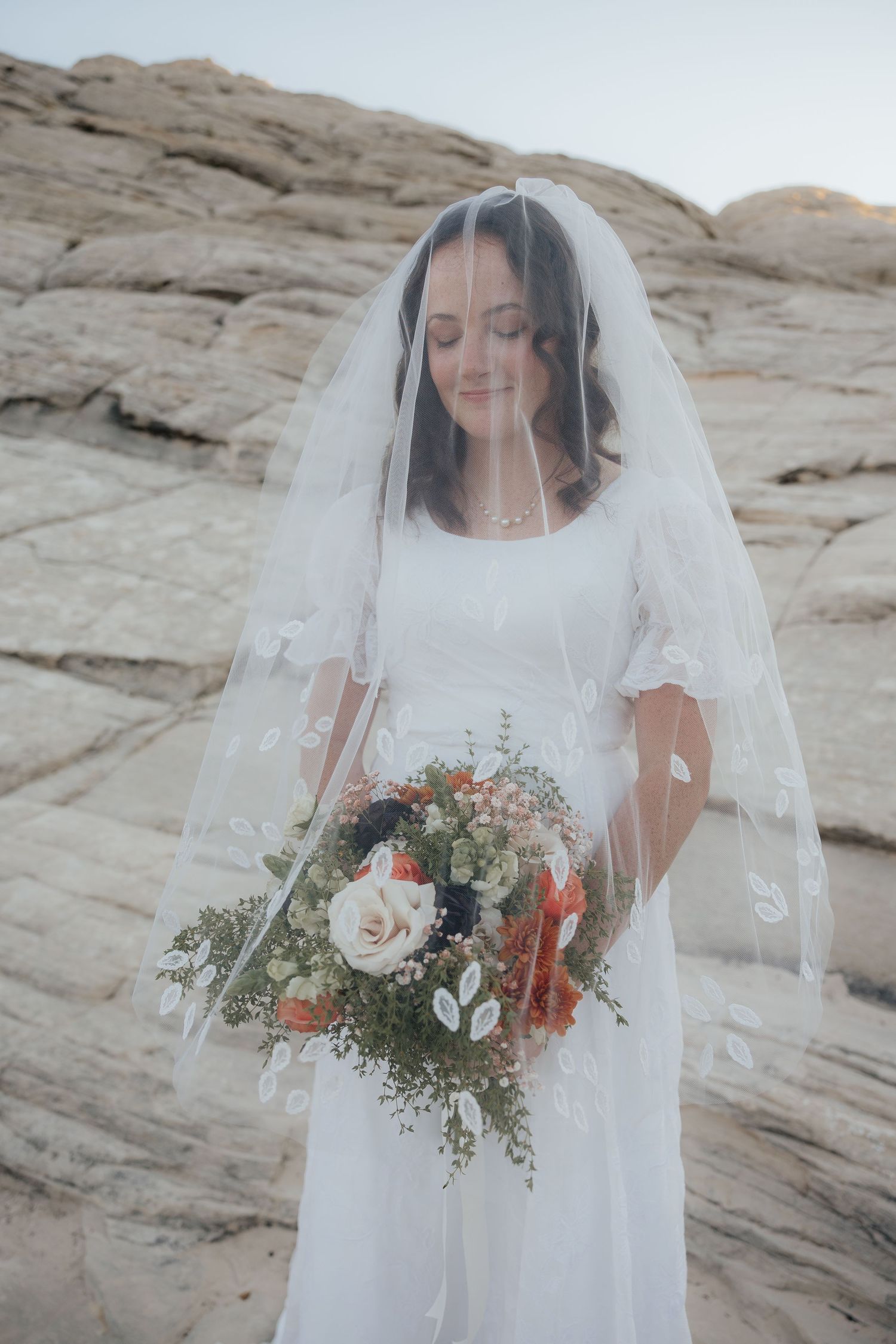 A bride in a white dress and veil holds a pastel bouquet while standing against rocky desert terrain.