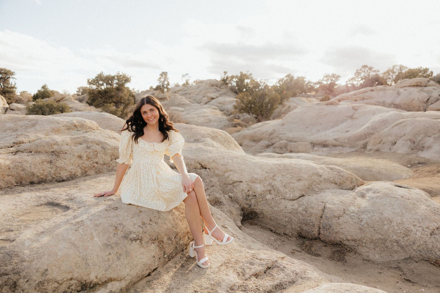 A figure in a flowing white dress sits gracefully on red rock formations in a desert landscape during golden hour.