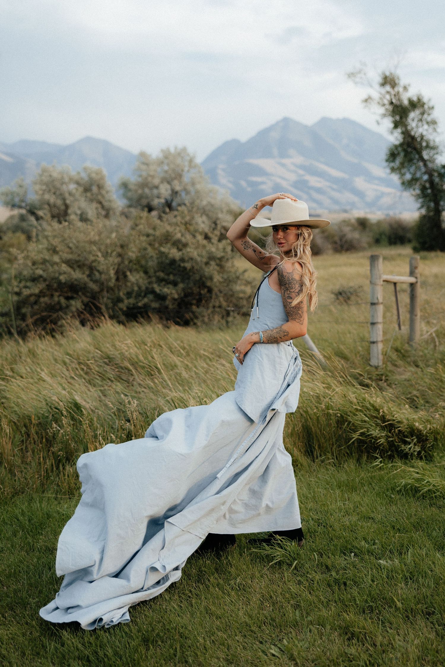 Tattooed woman in light blue gown and cowboy hat poses in mountain meadow with scenic peaks backdrop.