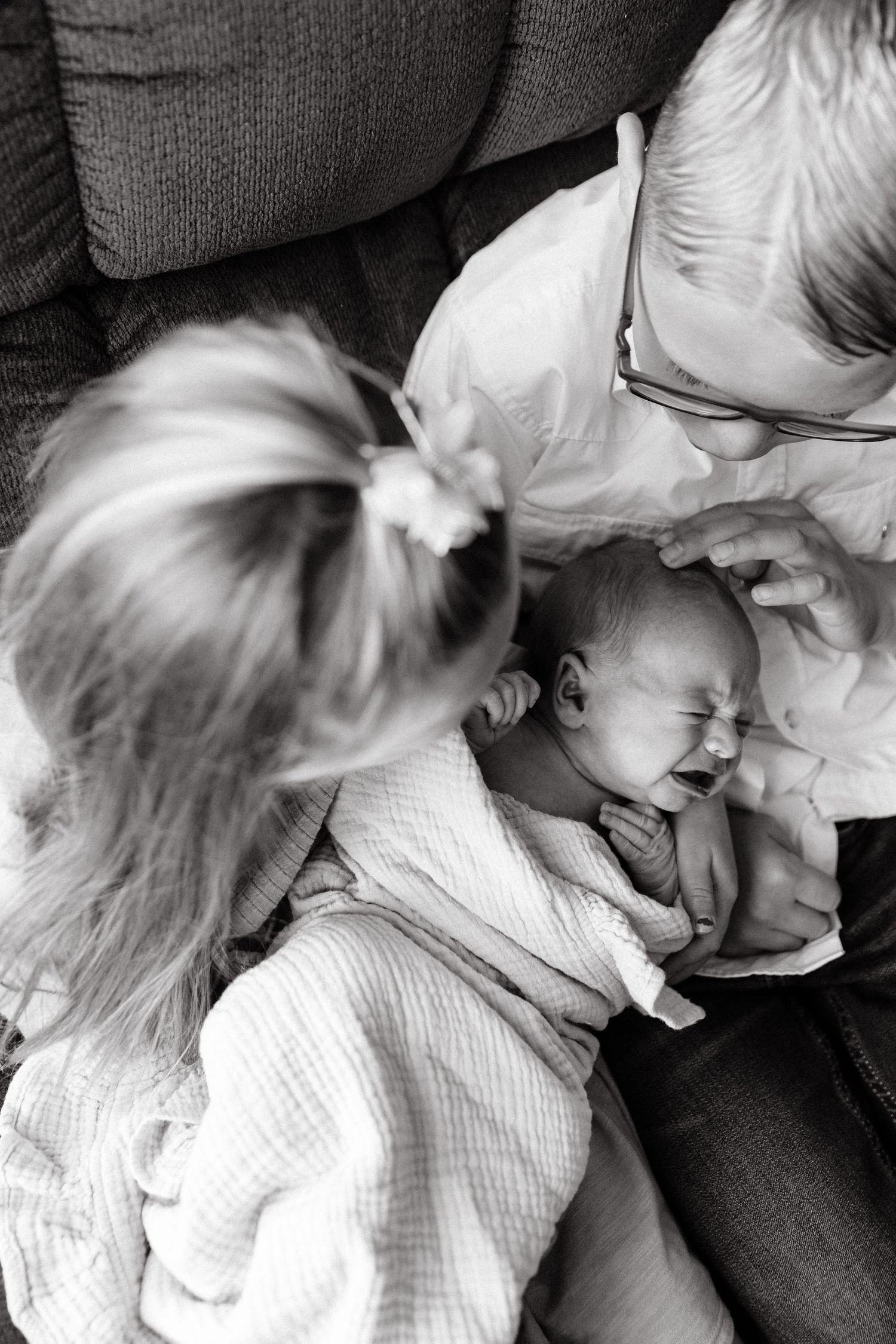 A black and white candid photo showing a tender moment as family members gather around a laughing infant on a couch.