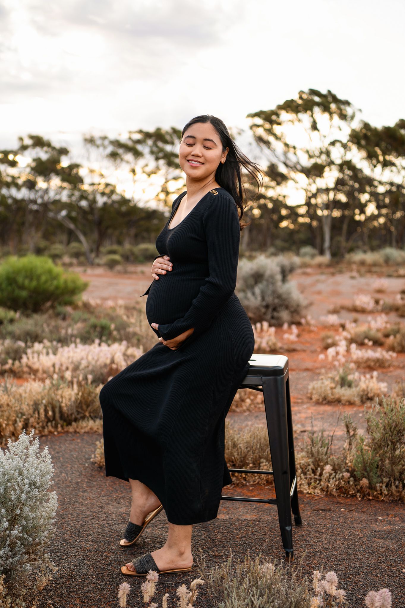 Pregnant woman in black dress sitting on stool outdoors in Australian bush landscape at sunset.