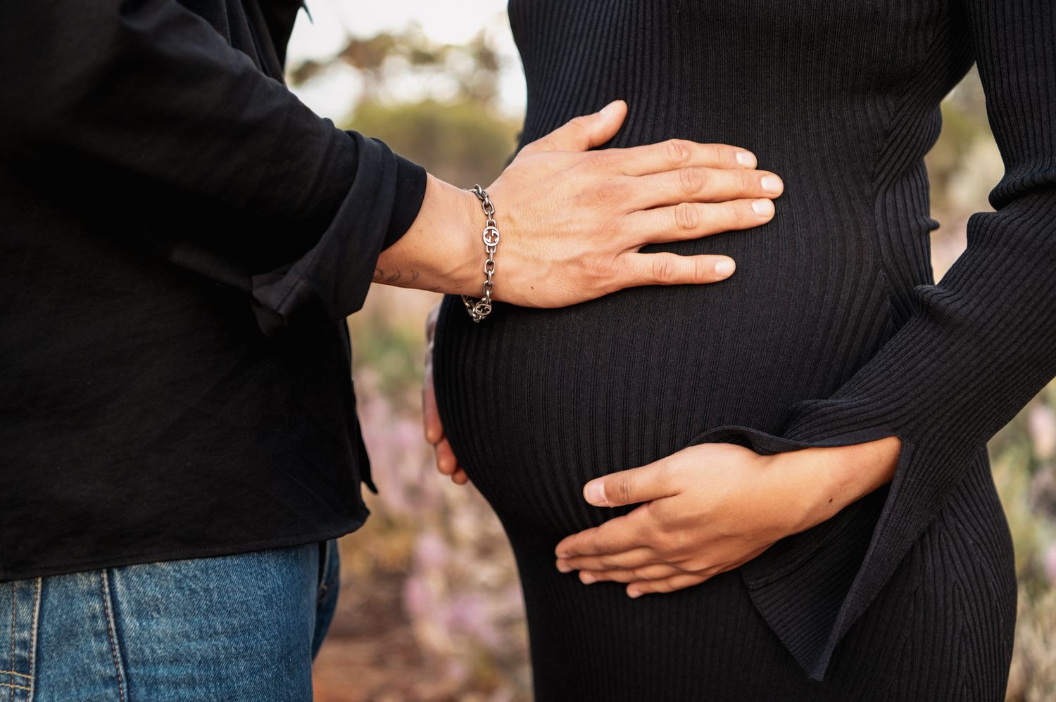 Couple tenderly cradling pregnant belly outdoors, woman in black ribbed dress with partner in jeans.
