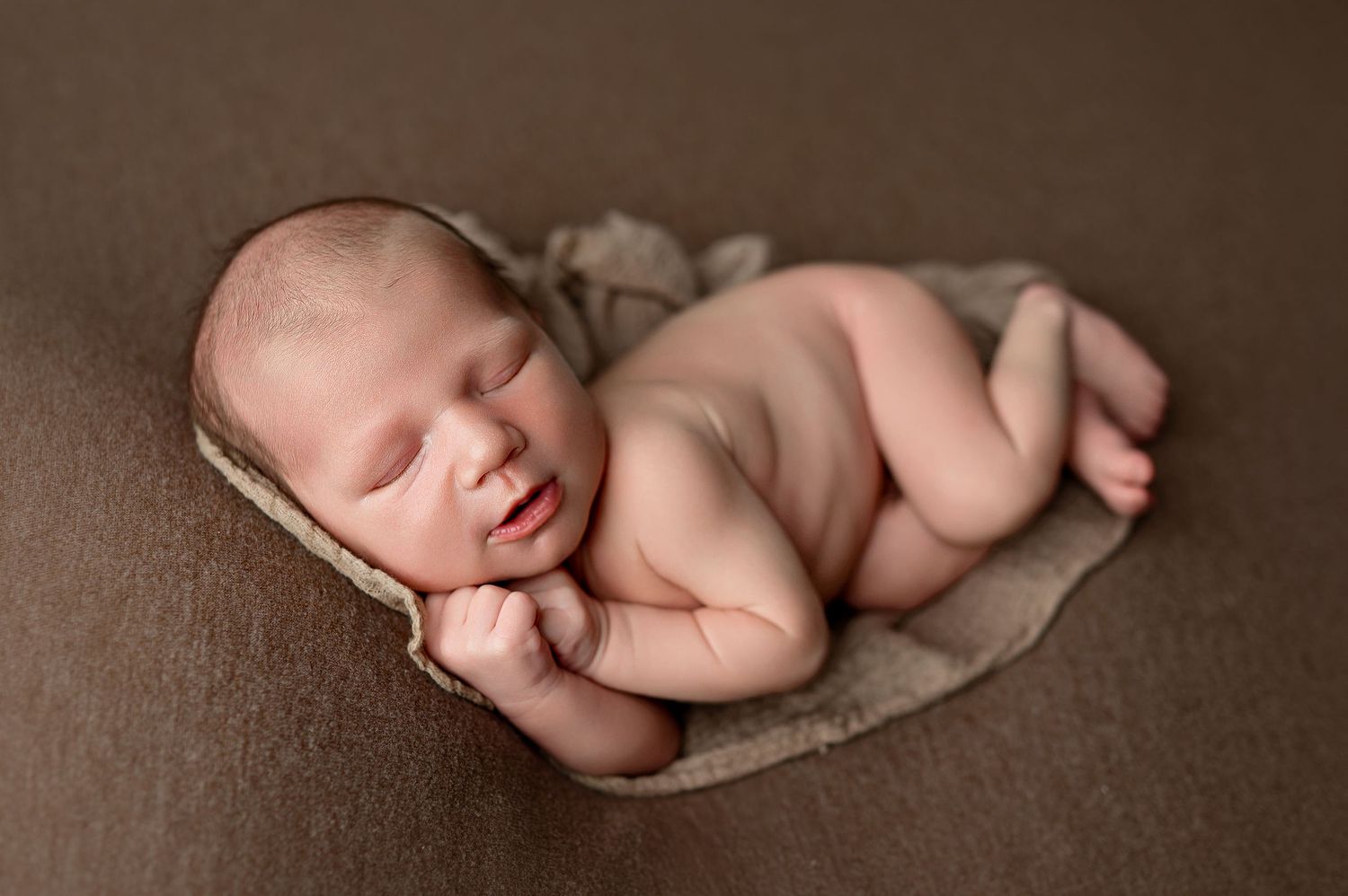 Peaceful newborn baby sleeping curled up on a brown blanket in a classic froggy pose during professional photo session.
