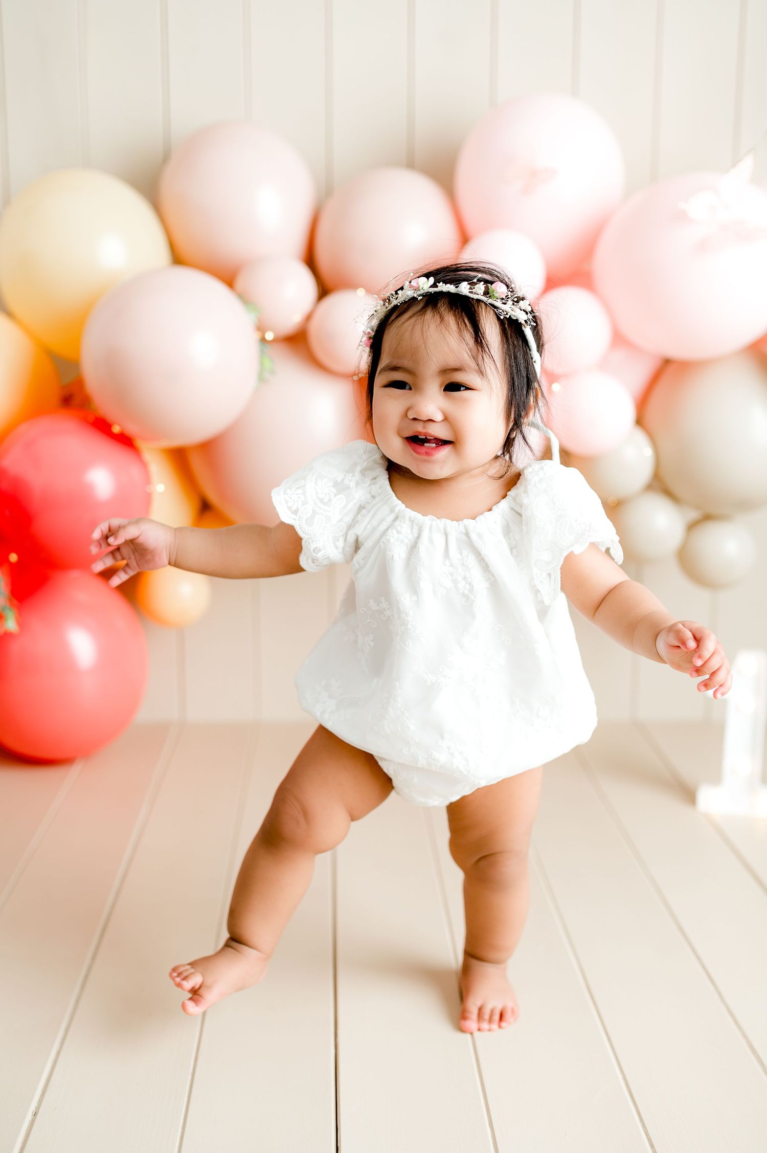 A baby in white dress sits and plays in front of a pink and peach balloon garland backdrop.