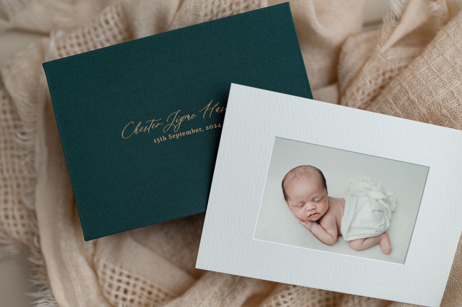 Professional photography studio album in dark green with newborn photo print displayed on textured fabric background.