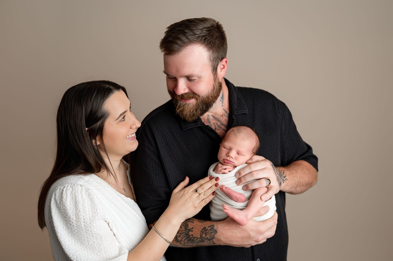 New parents smiling at their newborn baby, father holding infant while mother gently touches the baby in a studio portrait.