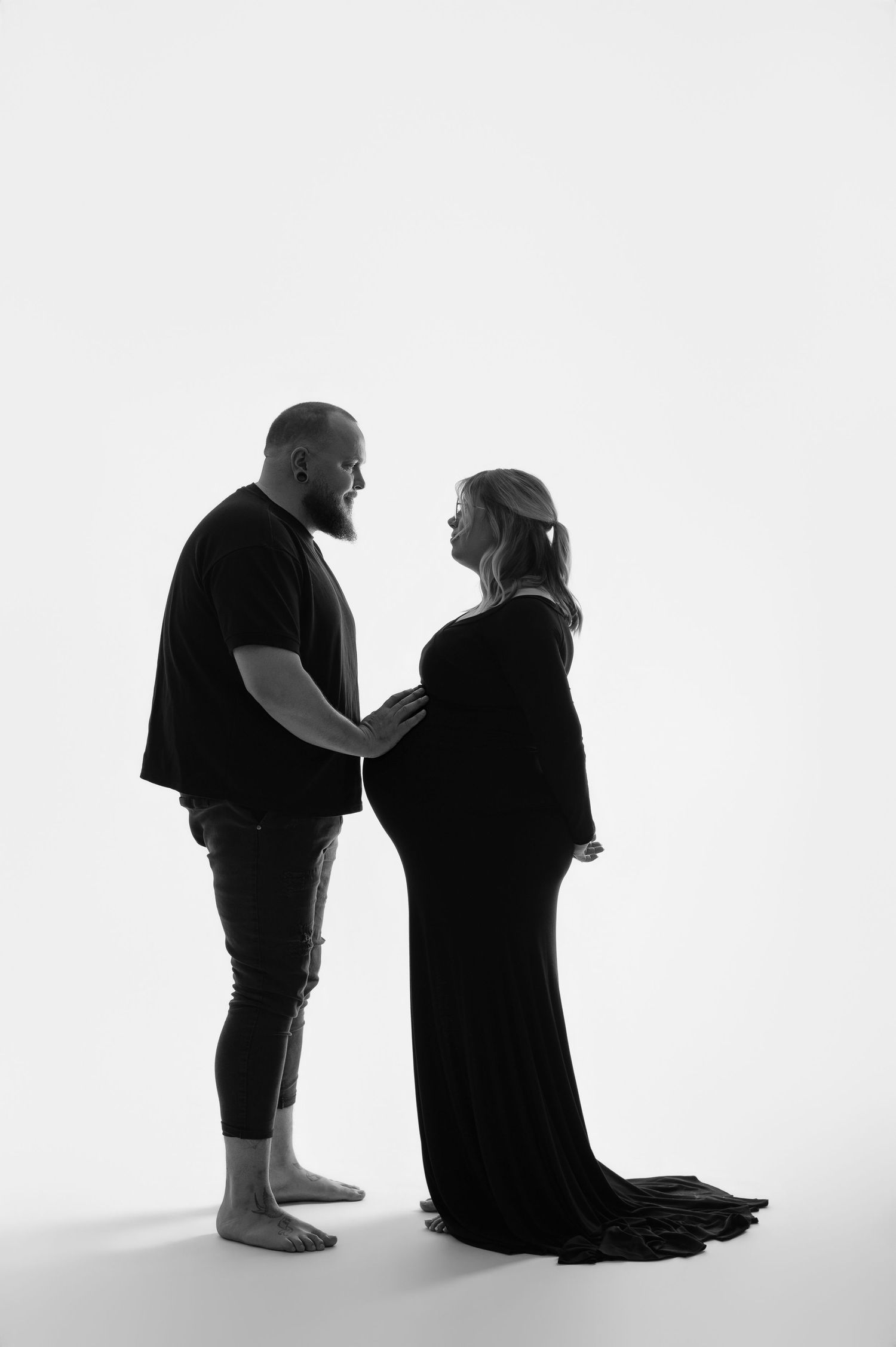 Black and white maternity photo of a couple in a studio, the pregnant woman in a flowing gown, both tenderly holding her belly.