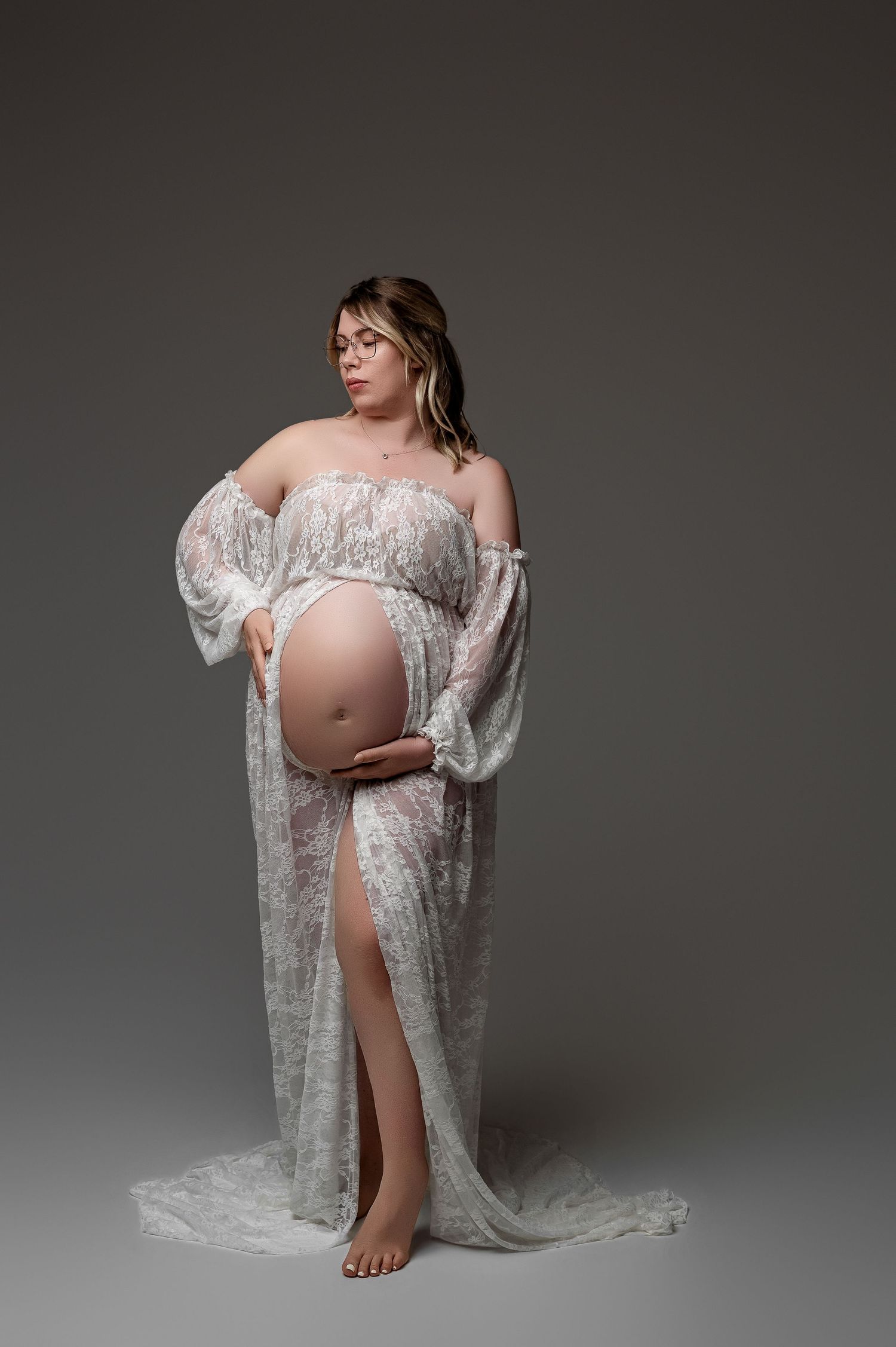 Pregnant woman in white lace maternity gown cradling baby bump, elegant studio portrait on gray background.