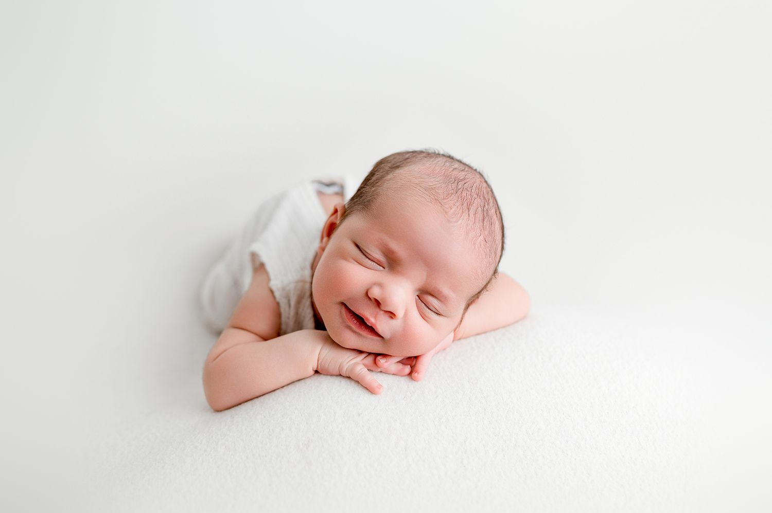 Smiling newborn baby in white romper lying on fluffy white blanket in professional studio portrait.