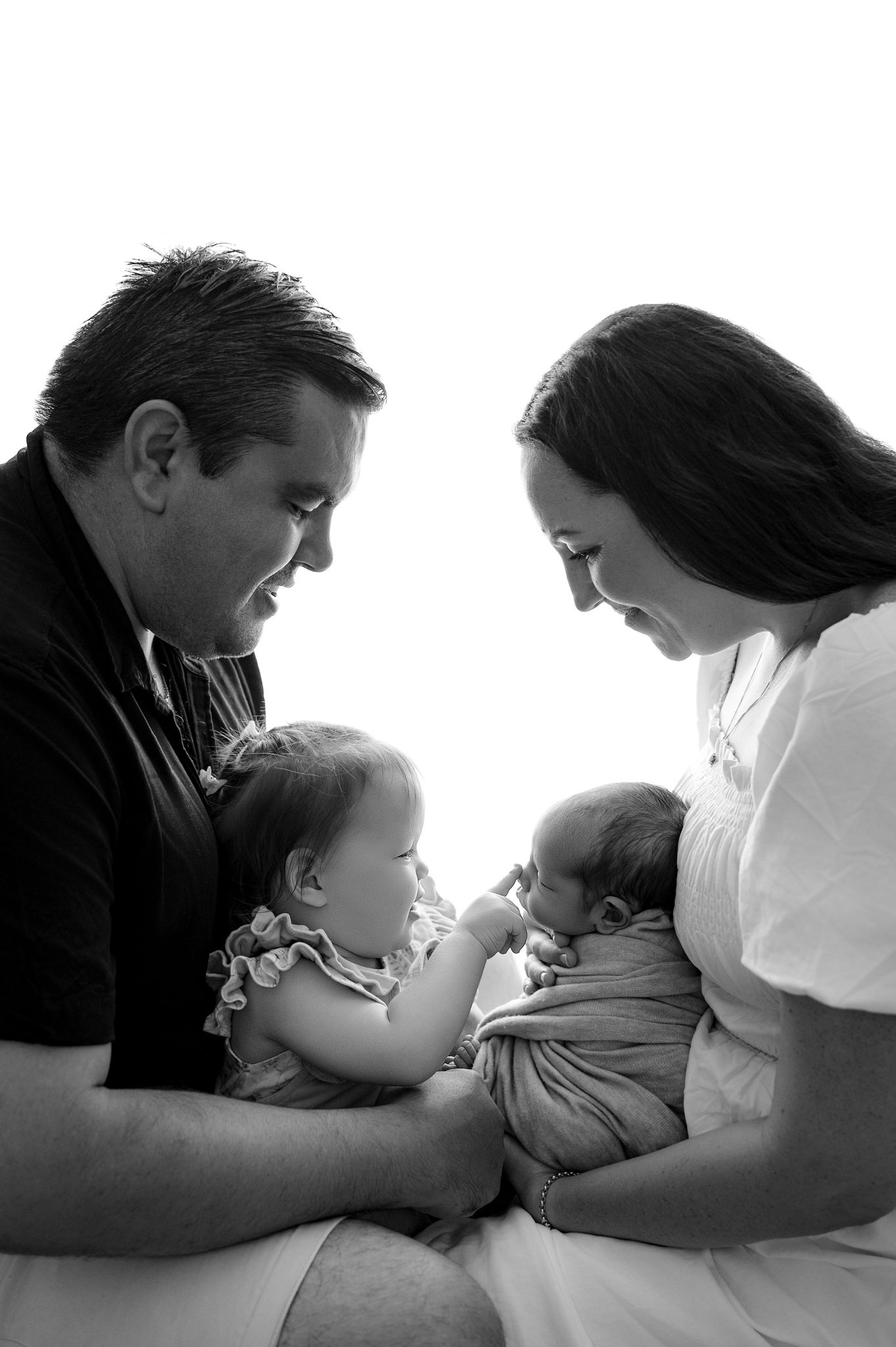Black and white family portrait of parents lovingly gazing at toddler and newborn baby.